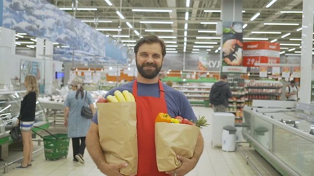 Handsome store employee in red aport with grocery paper bags standing in supermarket, he is smiling at camera