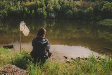 a young girl sits on the shore of a forest lake and catches fish in the early morning