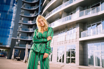 Beautiful girl in a green suit on a blurred building background. Stylish blonde girl. Fashionable girl. Everyday life of a beautiful girl. Lovely blonde in a stylish suit.