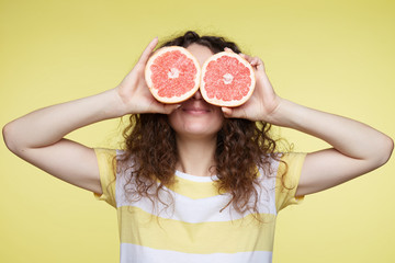 Isolated close up portrait of young curly haired female holding halved oranges at eyes. Headshot of funny girl wearing stripe T-shirt poses on yellow wall, smiles at camera. Expressions and emotions.