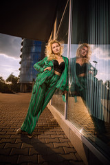 Naklejka premium A beautiful girl in a green suit stands near a glass wall at sunset in the city. Stylish blonde. The girl is reflected in the mirror surface. Everyday life of a beautiful girl. Beautiful blonde.