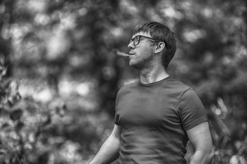 A young white bearded man in t-shirt and in glasses is vaping an electronic cigarette in the forest in the summer. Close up. Black and white.