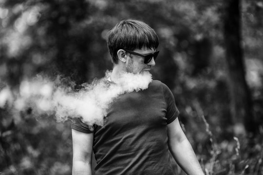 A young white bearded man in t-shirt and in sunglasses is vaping an electronic cigarette in the forest in the summer. Close up. Black and white.