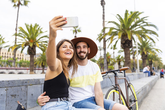 Spain, Barcelona, happy couple sitting on bench taking a selfie