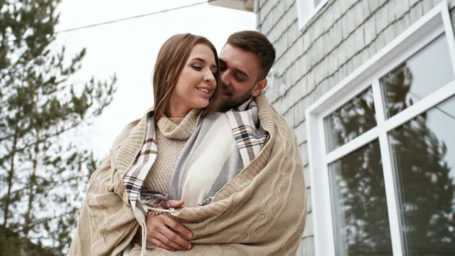 PAN with low angle of happy young couple wrapped in warm knitted blanket hugging and chatting while spending their morning on porch of forest house; pine trees in background