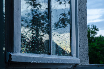 Window covered with raindrops after a summer rain, showing a reflection of the outside nature and sky basked in blue light of the evening