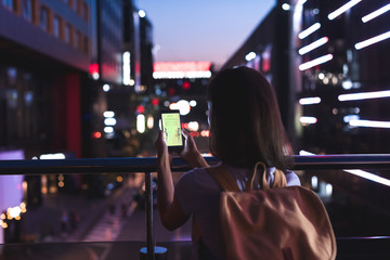 Fototapeta premium back view of woman with backpack and smartphone with shopping website on screen in hands standing on night city street
