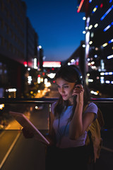 portrait of woman in headphones using tablet on street with night city lights on background