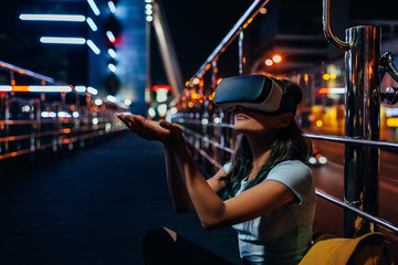 young woman in virtual reality headset sitting on street with night city on background