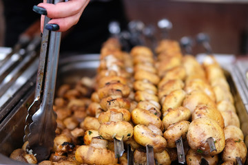 Grilled mushrooms on open kitchen food festival event. Street food and outdoor cooking concept. Hand of woman with thongs.
