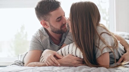 PAN of happy loving couple lying on bed and spending time together: they are rubbing noses and kissing gently