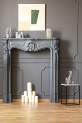Candles under fireplace portal in grey living room interior with poster and table. Real photo
