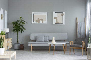 Posters above grey sofa in minimal living room interior with plants and armchair. Real photo