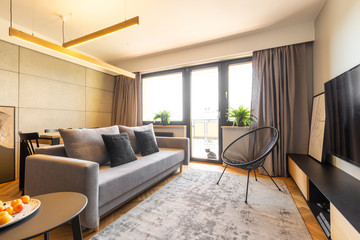 Armchair on carpet and grey sofa in bright living room interior with balcony. Real photo