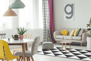 Real photo of a creative living room interior with a poster on the wall, couch, pouf, dining table, chairs, and geometrical carpet