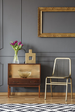 Gold chair next to wooden cabinet with flowers in grey interior with mockup. Real photo