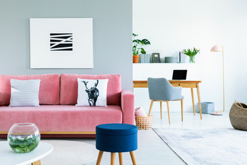 Poster above pink sofa with pillows in open space interior with blue stool and armchair. Real photo