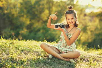 Child girl sitting on the grass. In her hands she has an old photo camera and she shows gesture all...