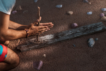 A man tries to get fire with an ancient method. Sandy beach in the background - obrazy, fototapety, plakaty
