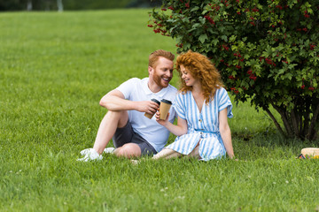 Fototapeta premium happy redhead couple clinking by paper cups with coffee on grass in park