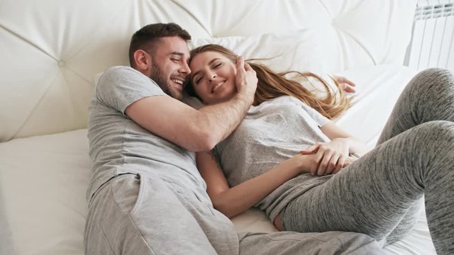 Handheld shot happy bearded man talking and caressing face of beautiful girlfriend lying on bed next to him and laughing
