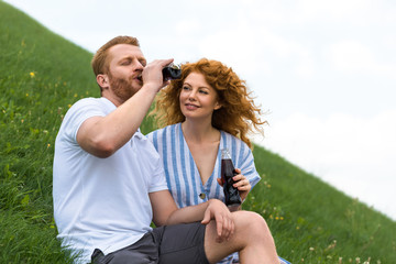 redhead man with closed eyes drinking soda from bottle near smiling girlfriend on grassy hill