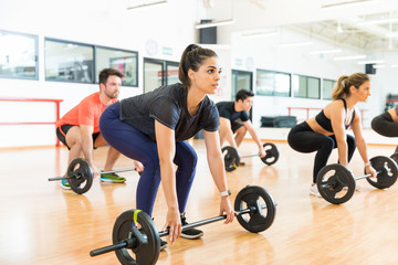 Woman Picking Up Barbell While Exercising With Friends In Gym