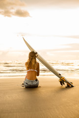 Junge Surferfrau im sexy weißen Bikini mit leerem surfendem Shortboard sitzen auf Sand am Sonnenuntergangozean. Moderner Familienlebensstil, Wassersport-Abenteuercamp für Menschen und extremes Schwimmen im Sommerurlaub. © Mikalai Bachkou