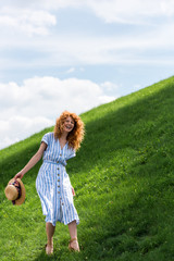 happy beautiful redhead woman with straw hat in hand standing on grassy hill against blue sky