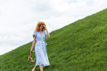 attractive redhead woman carrying shoes on grassy hill