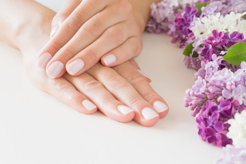 Fototapeta premium Young, perfect woman's hands with light nails on pastel pink table. Care about clean, soft, smooth skin. Manicure, pedicure beauty salon. Beautiful branches of lilac blossoms. Colorful, fresh flowers.