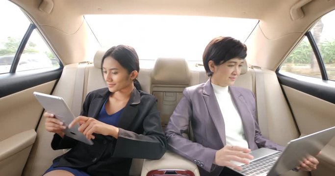 Two young businesswomen with tablet computer and laptop in the backseat of a car.