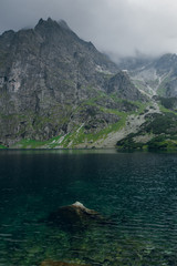 Scenic view of foggy mountains cover by dark clouds and green forest with a reflection in a lake. Stony shore. Morskie Oko. Marine Eye. High Tatras, Zakopane, Poland
