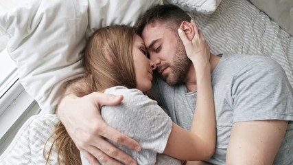 Handheld shot of affectionate young couple lying in bed and touching their foreheads together while hugging and sharing intimate moment