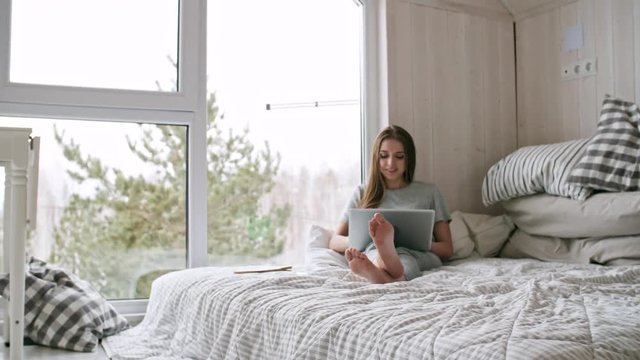 PAN of cheerful young woman sitting on bed and talking to someone on video call on laptop