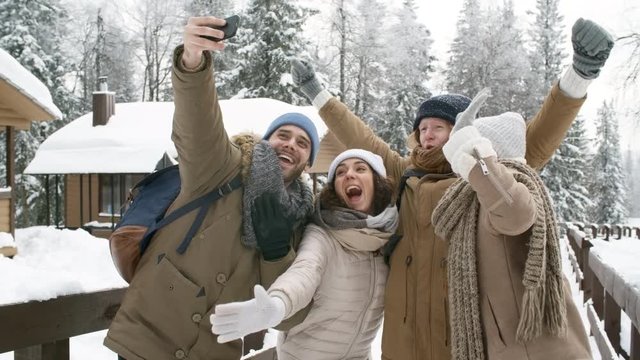 Group of excited young friends waving and smiling at camera while taking selfie with smartphone in winter forest