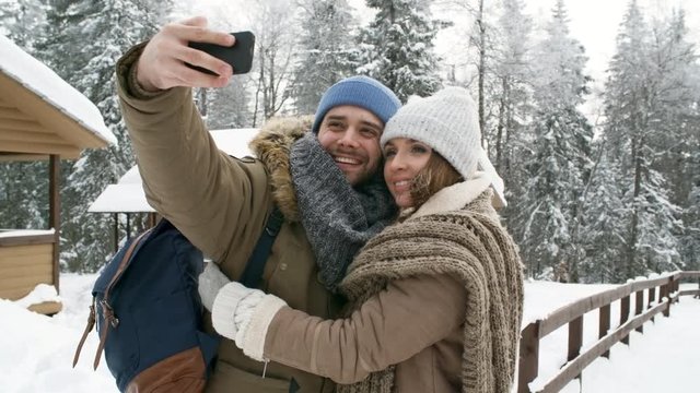 Beautiful young couple smiling and embracing while taking selfie with smartphone in forest at snowy winter day
