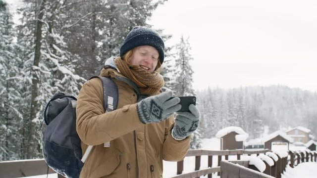 Happy young redhead guy smiling at camera of smartphone and taking selfie in the park at snowy winter day