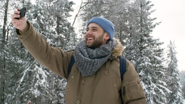 Joyful handsome man video calling on smartphone, waving at camera, smiling and telling about beautiful winter nature in national park while walking at snowy day