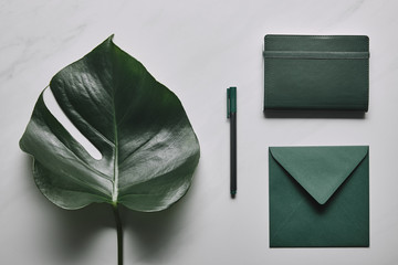 Green envelope and notebook with pen on white marble background with monstera leaf
