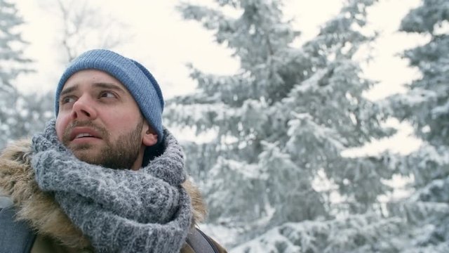 Close-up shot of freezing male tourist in knitted scarf and hat looking from side to side in snowy forest while hiking at winter day