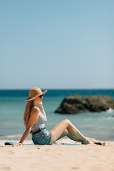 Side view an attractive young woman wearing straw hat and sunglesses sits on a beach with ocean background