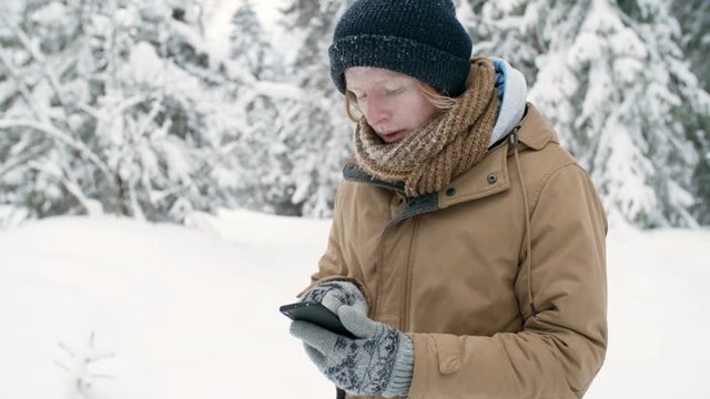 Young redhead guy standing in snowy forest and using navigator on smartphone during winter hike