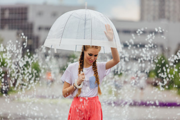 Young pretty girl with two braids in yellow boots and with transparent umbrella stands near fountain.