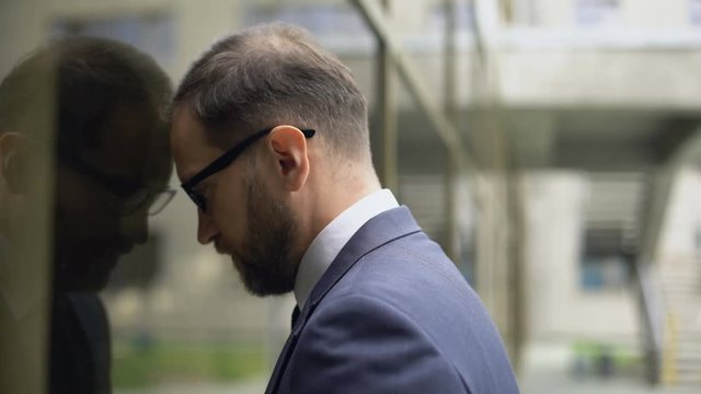 Exhausted office worker in suit leaning on glass building wall, depression