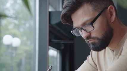 Man drinks a morning coffee in a light coffee shop and makes notes in a notebook. Modern man sits at the window in a cafe and writes in an organizer or a diary. Coffee and croissant on the table.