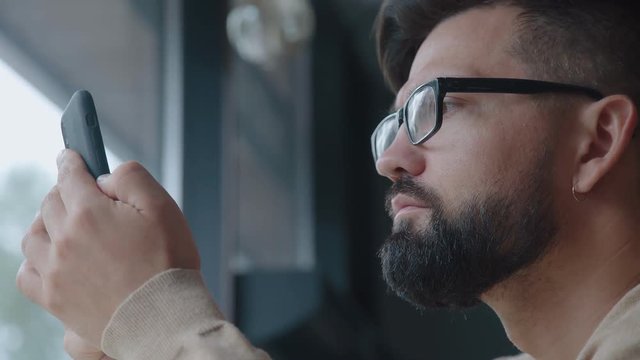A man in glasses holds a phone in front of his face and looks at the screen (reading news, social networks). A man with a beard and a smartphone: he looks at the screen, swype of the page.