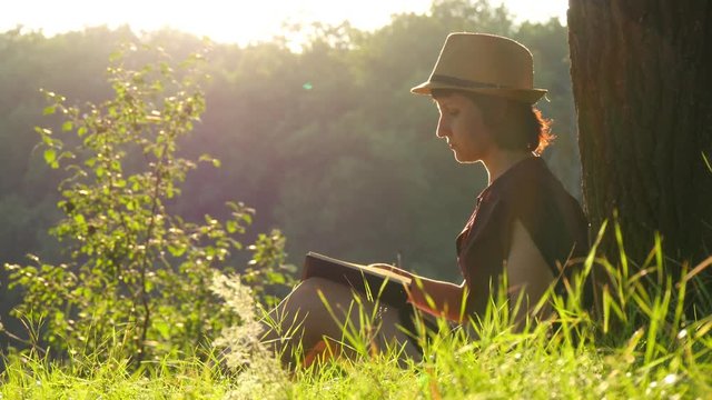 A young female student is sitting in a Park under a tree and reading a book. Education and reading outdoors.