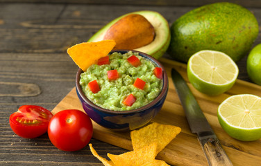 Guacamole in blue bowl on natural rustic desk with ingredient: lemon, tomatoes, peppers around.