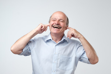 Good-looking happy mature european man smiling with closed eyes, holding index fingers in ears, waiting for surprise